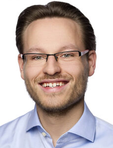 ein junger Mann mit kurzen dunklen Haaren und Brille. Er trägt ein blaues Hamd und lächelt. 