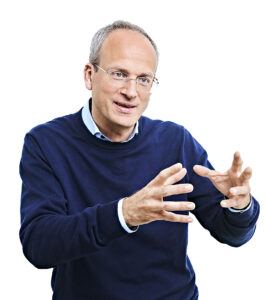 Eon Mann mit zurückweichendem Haaransatz und Brille. Er trägt einen dunkelblauen Puli über einem hellen Hemd und hält beide Hände gestikulierend vor sich.