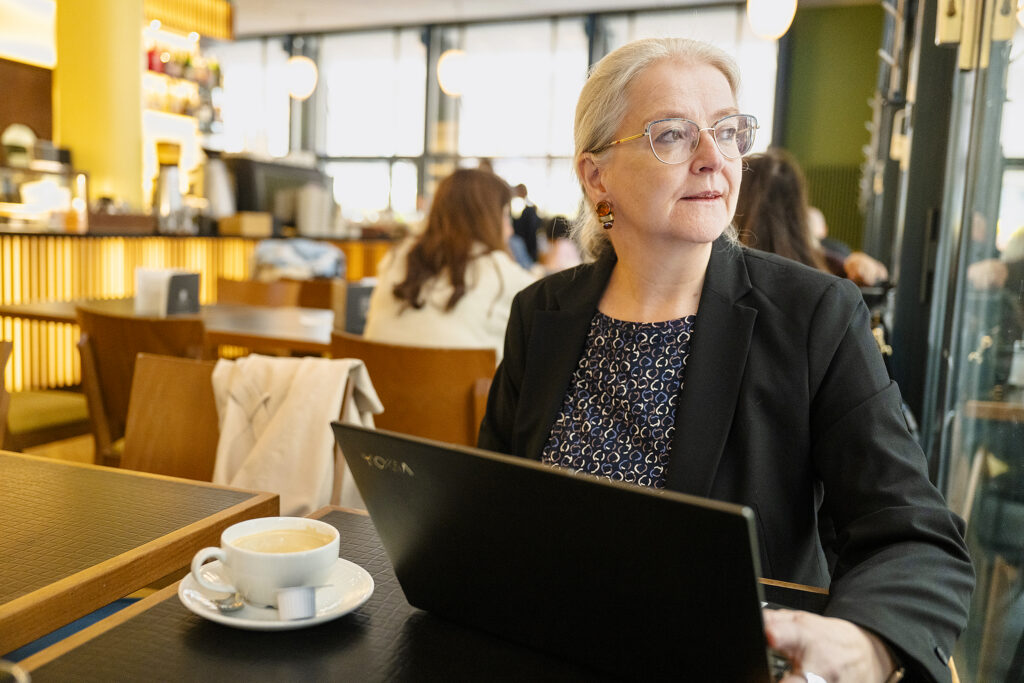 Eine Frau mit zurückgebundenen blonden Haaren und Brille sitzt mit ihrem Laptop in einem Café. Sie schaut sinnierend zur Seite.