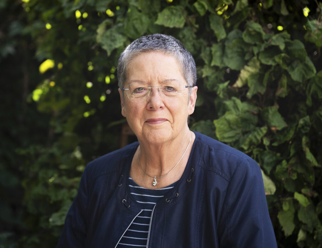 Eine Frau mit sehr kurzen grau-melierten Haaren und Brille. Sie seht vor einem grünen Busch und lächelt in die Kamera.