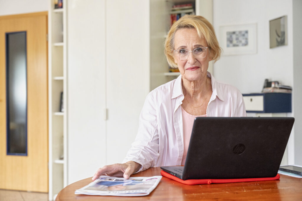 Eine Frau mit blonden halblangen Haaren sitzt hiinter einem Laptop in einem Büro. Sie lächelt in die Kamera.