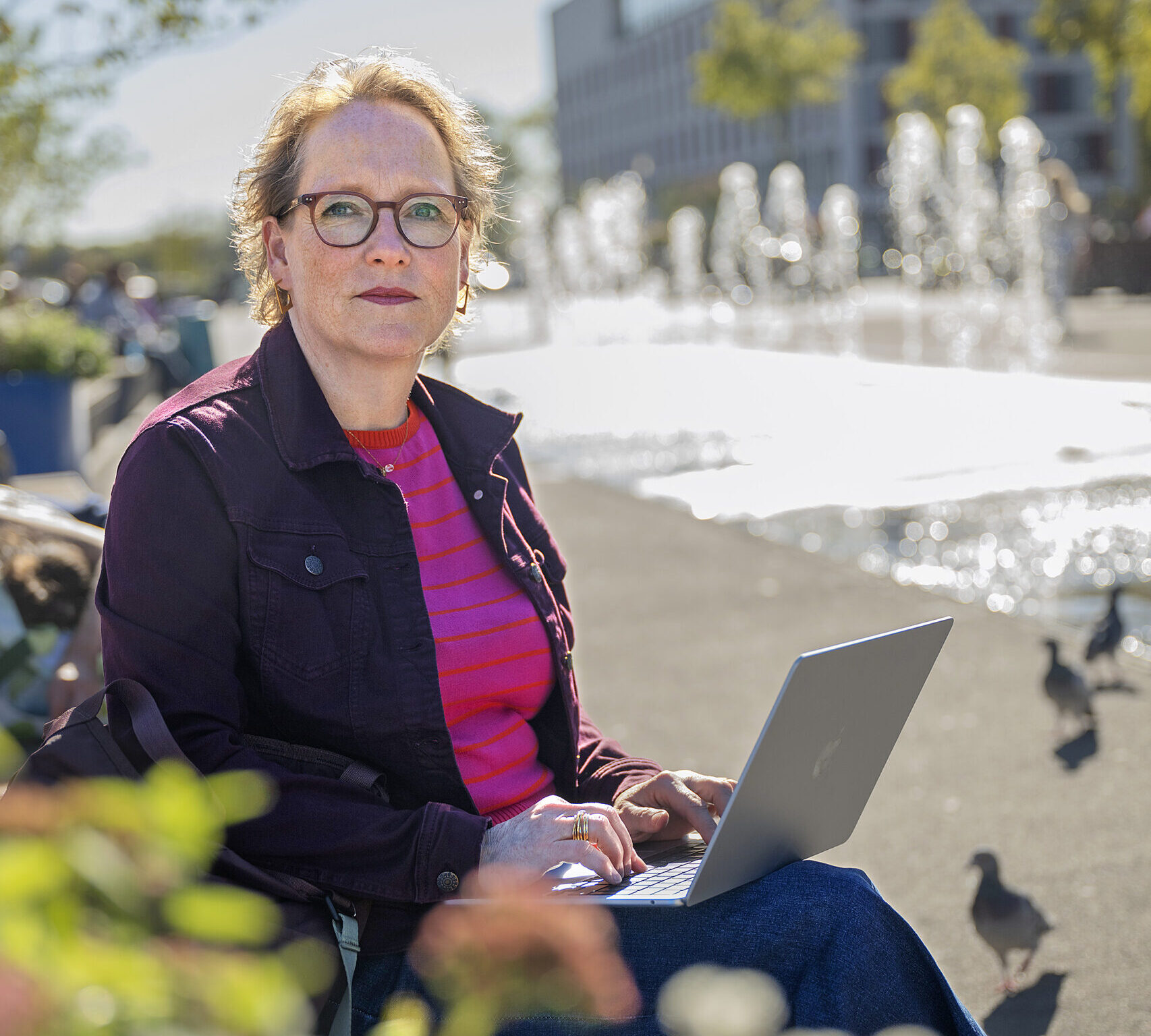 Eine Frau mit rötlichen Haaren und Brille sitzt mit einem Laptop auf dem Schoß im Freien. Im Hintergrund ist unscharf ein Brunnen zu erkennen.